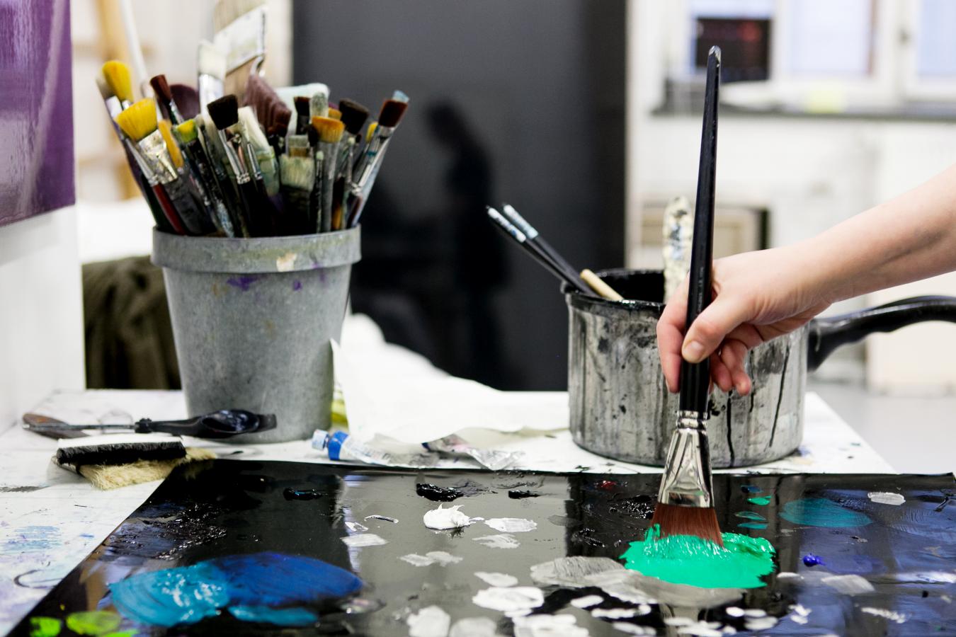Kuvassa näkyy taiteilijan käsi, joka maalaa pensselillä taulua, sekä taustalla taiteilijan työvälineitä.