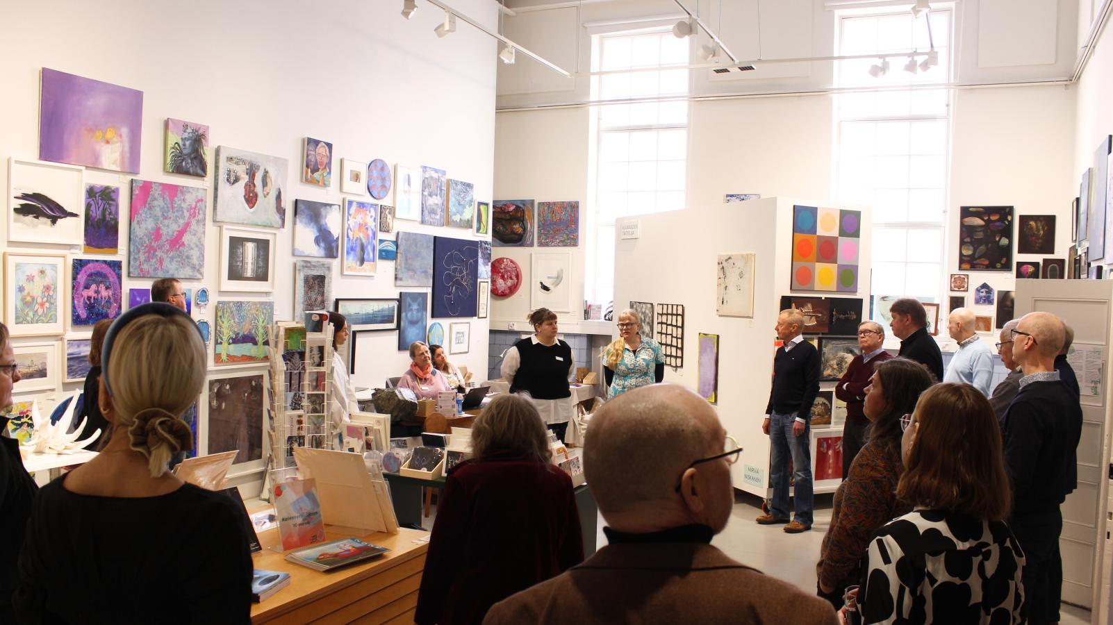 Kuvassa näkyy vaalea näyttelytila, jonka seinillä on paljon eri kokoisia ja värisiä maalauksia. Tilaan on kokoontunut joukko ihmisiä