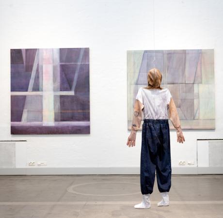 Maalauksia museotilassa, maalausten edessä roikkuu ihmisen hahmoa mukaileva teos